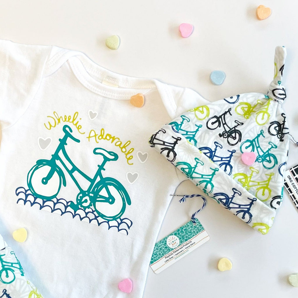 A bicycle themed baby gift set that includes an organic cotton baby onesie printed with a turquoise bike that says "Wheelie Adorable" in chartreuse green and a coordinated organic cotton newborn baby hat printed with blue/green bicycles.