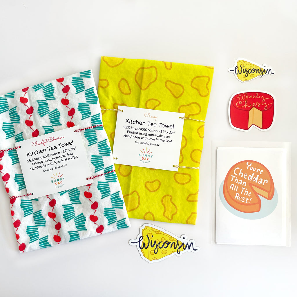 a Wisconsin-themed Gift Set of 6 products is shown on a white background. Featuring a cherry printed kitchen tea towel, a cheese printed tea towel, a Wisconsin cheese-themed vinyl magnet, a WI cheese vinyl sticker, a "Wheely Cheesy" cheese wheel pun vinyl sticker & a cheese-themed greeting card that says "You're Cheddar Than All The Rest".