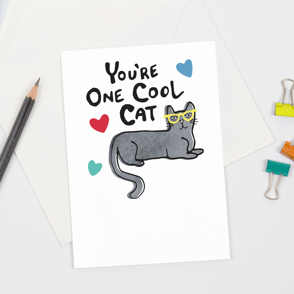 A cat-themed appreciation greeting card is shown on a white background next to a pencil and colorful binder clips - the cover of the greeting card shows a gray watercolor cat wearing yellow glasses, red, green, and blue hearts, and hand lettered black text that says "You're One Cool Cat"
