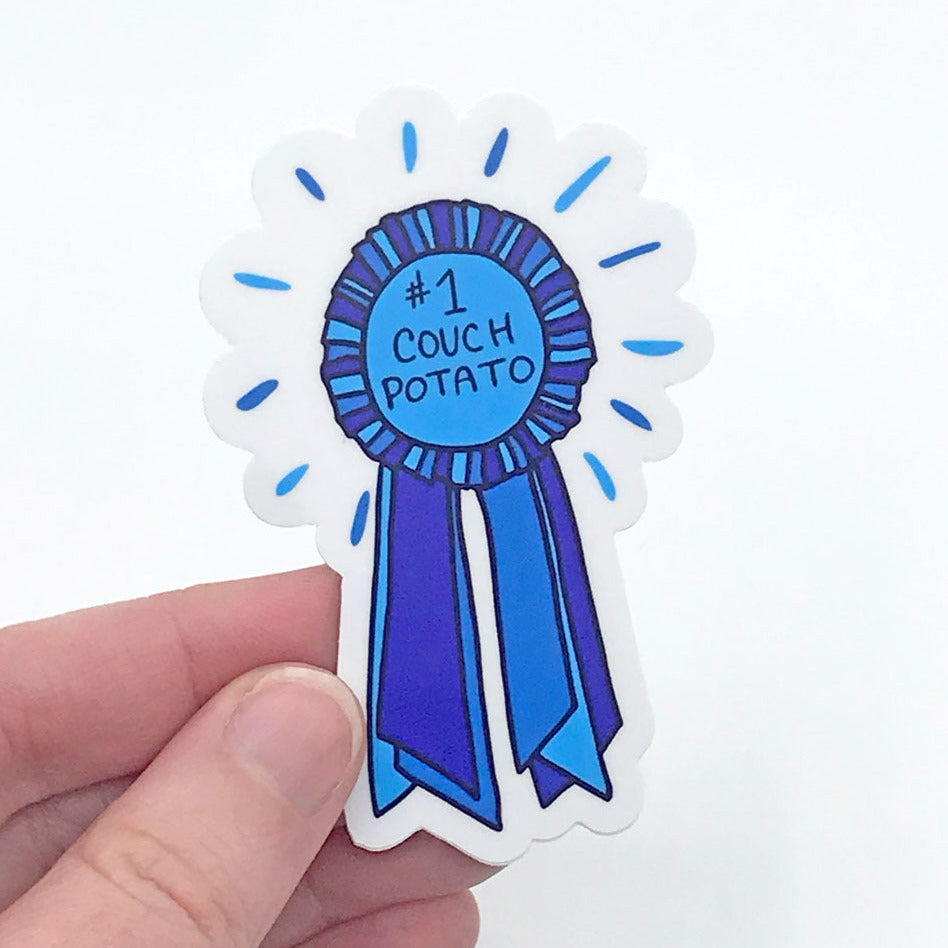 A fun vinyl sticker design being held in a hand against a white background. Featuring a 1st place, blue ribbon award with the words "#1 Couch Potato", this illustrated vinyl sticker by Sunny Day Designs makes a funny gift for anyone who loves being lazy! Made in the USA.