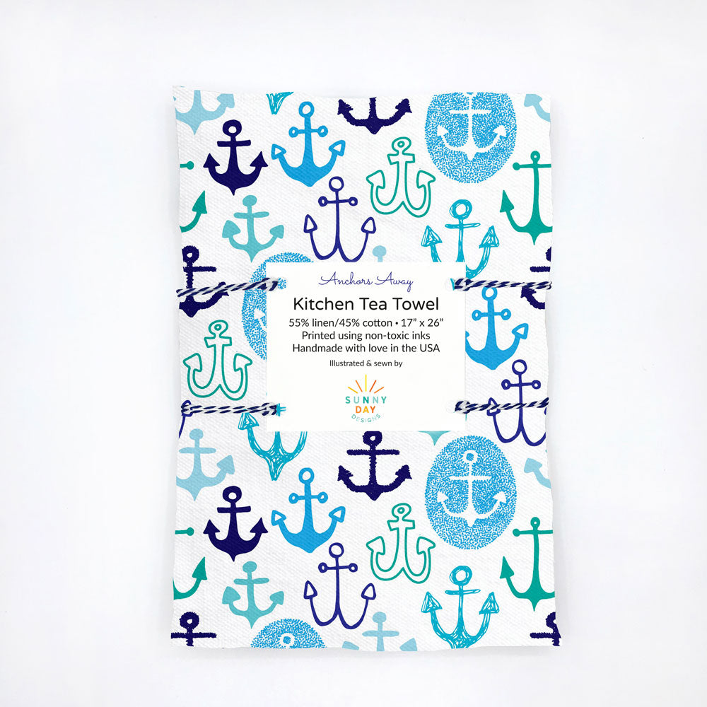 Whimsically hand drawn anchor shapes in shades of blue, turquoise, and navy are scattered across a white background. This image shows our printed "Anchors Away" kitchen tea towel design by Sunny Day Designs. The tea towel lis folded and packaged with a descriptive postcard and wrapped in blue striped baker's twine.