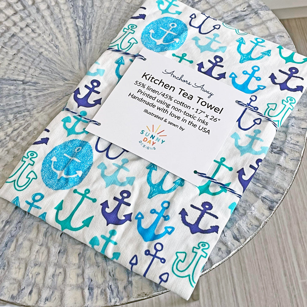 Whimsically hand drawn anchor shapes in shades of blue, turquoise, and navy are scattered across a white background. This image shows our printed "Anchors Away" kitchen tea towel design by Sunny Day Designs. The tea towel lis folded and packaged with a descriptive postcard and wrapped in blue striped baker's twine.