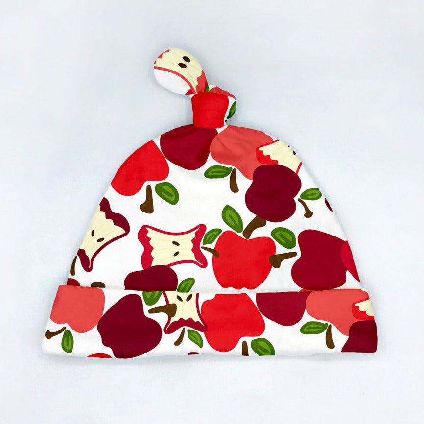 A red apple printed organic cotton baby hat by Sunny Day Designs is pictured on a white background. This cute fruit-themed newborn hat is perfect for Fall, made in the USA, and features a tie top style.