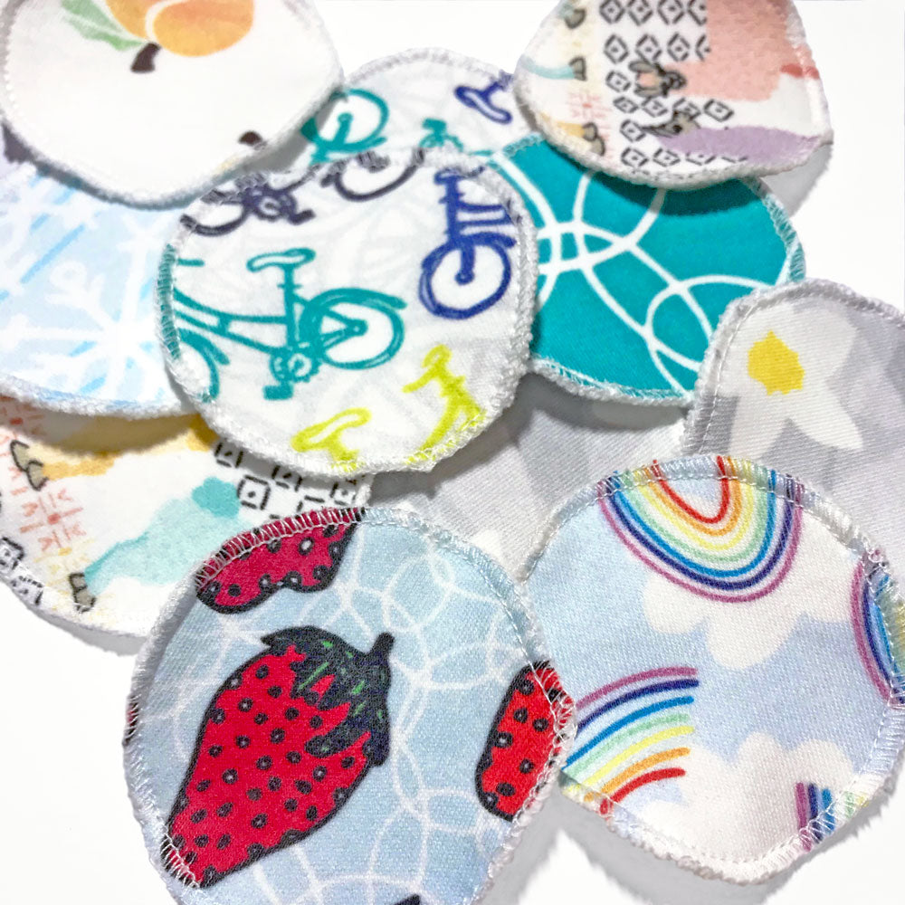 A selection of colorful, organic cotton printed reusable facial rounds made in the USA by Sunny Day Designs and scattered on a white background.