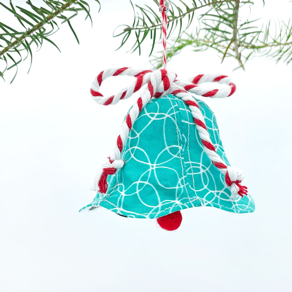 This colorful and cute handmade Christmas ornament is made from stitched turquoise/white fabric by Sunny Day Designs and trimmed with red/white baker's twine and a red wool pom pom. Holiday ornament is shown against a white background with ornament hanging from Christmas tree branches.