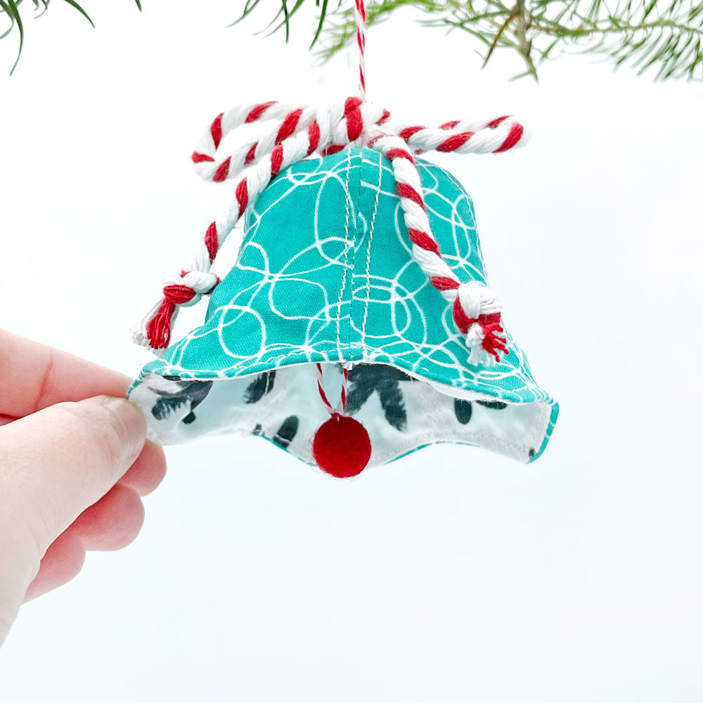 A handheld view of our handmade Christmas Bell ornament with the bell being tipped up to show the contrasting fabric on the inside of the bell. This holiday ornament is shown with the contrasting black/white fabric visible against the turquoise/white fabric on the front of the bell. Trimmed with red/white cotton twine bow and a red pom pom.