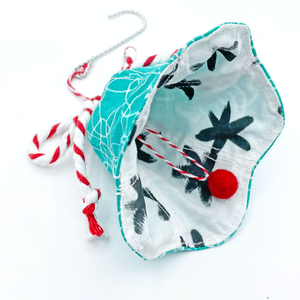 This bell shaped Holiday ornament by Sunny Day Designs is turquoise, white, black, and red. Made in the USA by Sunny Day Designs out of fabric.