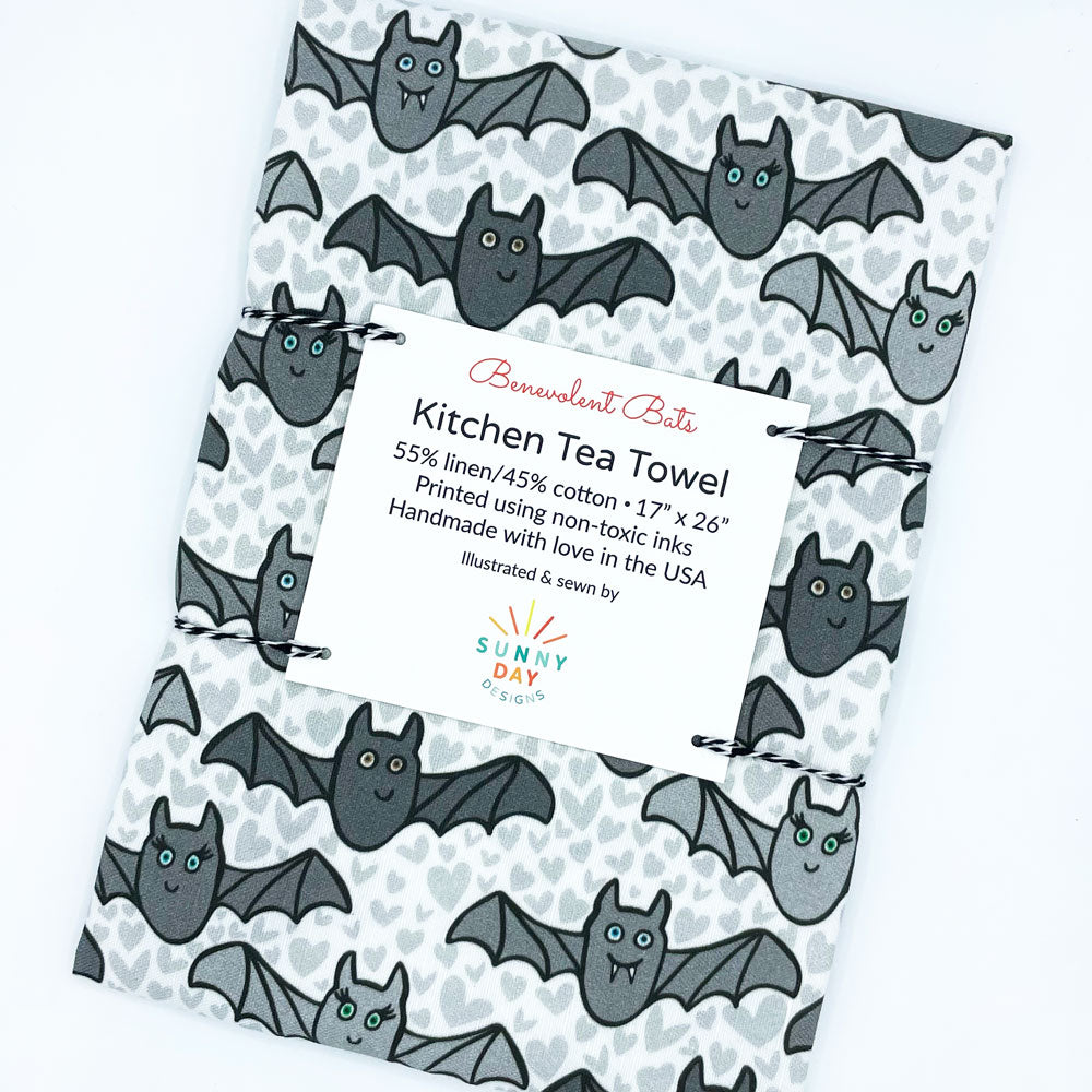 Our cute and whimsical Benevolent Bats printed kitchen towel is perfect for decorating kitchens for Halloween! This fun tea towel by Sunny Day Designs will make you smile, and you'll love that each dish towel is made in the USA from linen/cotton fabric. This image shows the towel packaged and slightly tilted on a white background.