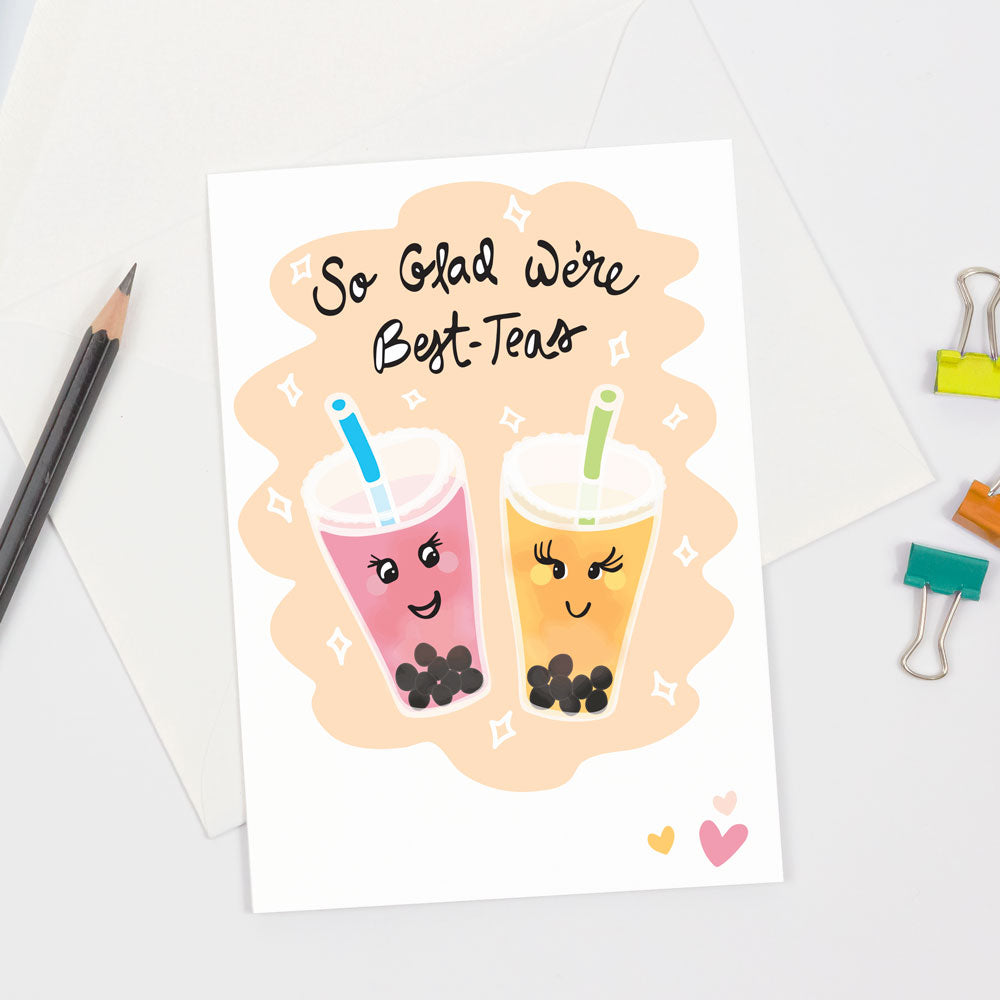 Colorful bubble tea-themed greeting card shown on a white table with colorful binder clips and a pencil. The greeting card is the "Best-Teas" Friendship greeting card by Sunny Day Designs, which is printed on sustainably sourced paper and features 2 colorful, smiling bubble tea drinks on a tan and white background with 3 hearts in the bottom right corner. The hand lettered  text on the front of the card reads "So Glad We're Best-Teas"