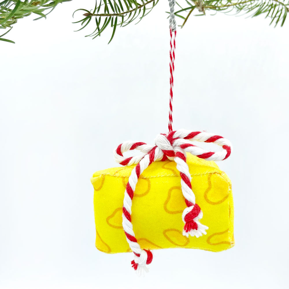 3D handmade yellow Cheese Christmas Ornament is made from 100% organic cotton fabric and features a Red and white striped baker's Twine Bow at the top. This holiday ornment is made in the USA by Sunny Day Designs and the image shows it hanging from a Christmas treea branch against a white background.