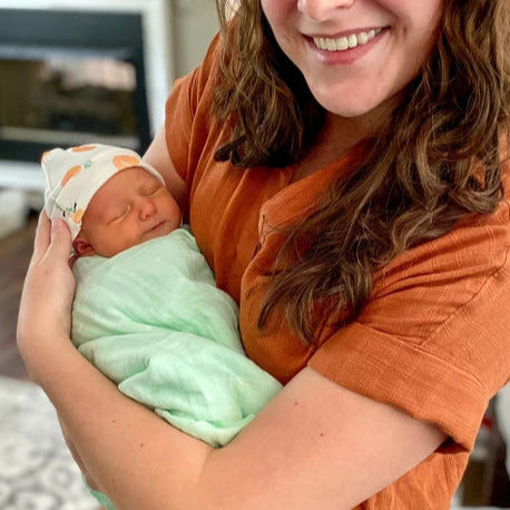 This is a customer submitted image showing a newborn baby wearing a orange, green, and white organic cotton baby hat in our Peachy Keen peach fruit print design and a green blanket. In the image, the customer is holding the sleeping newborn baby while it wears the Peachy Keen handmade baby hat by Sunny Day Designs.
