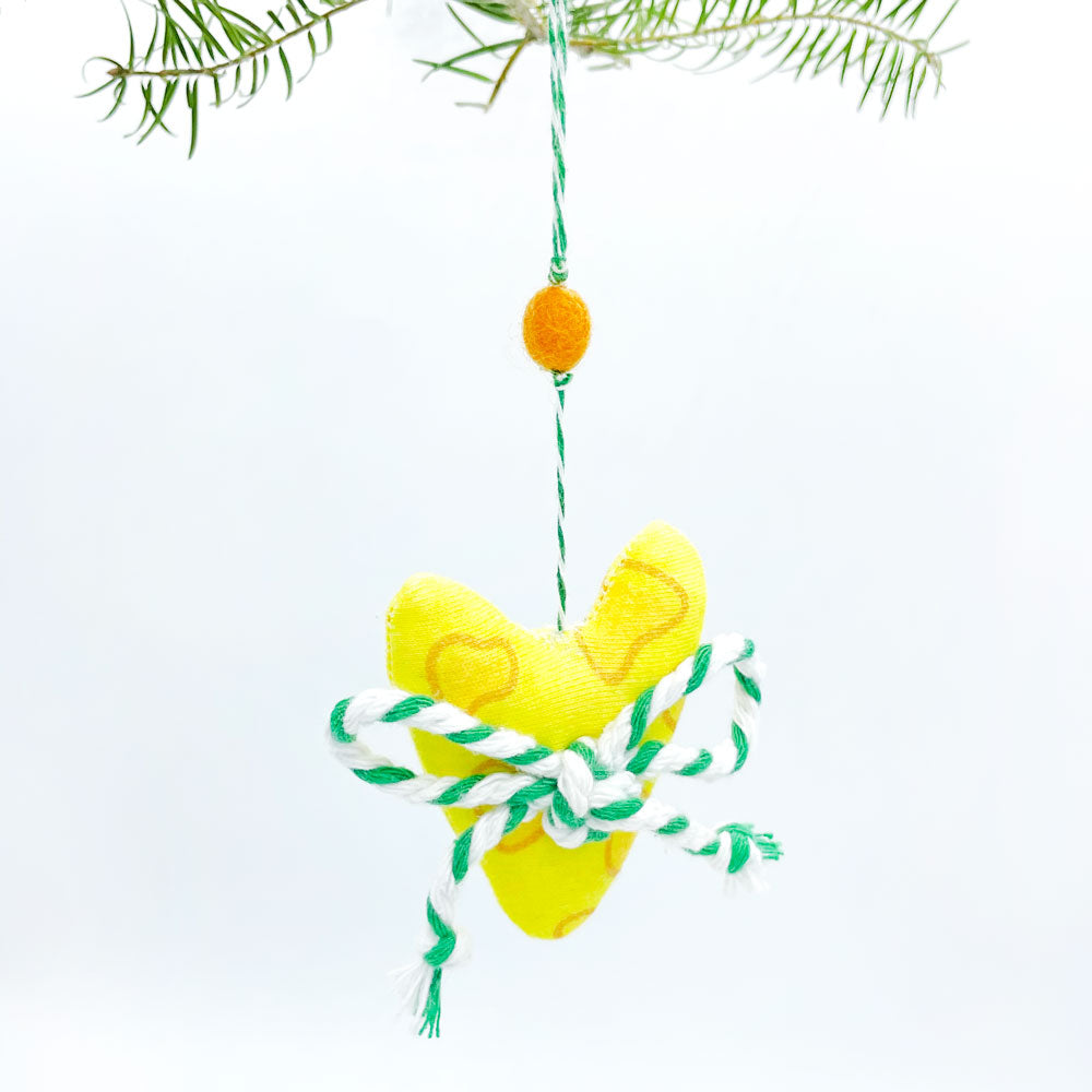A cute heart shaped Christmas ornament made from cheesy printed, organic cotton fabric and topped with a green/white baker's twine bow hangs from Christmas tree branches against a white background. This handmade holiday ornament is made in Wisconsin by Sunny Day Designs and is topped with a mustard yellow wool pom pom. This ornament is a great gift for Green Bay Packers fans and is also made in America.