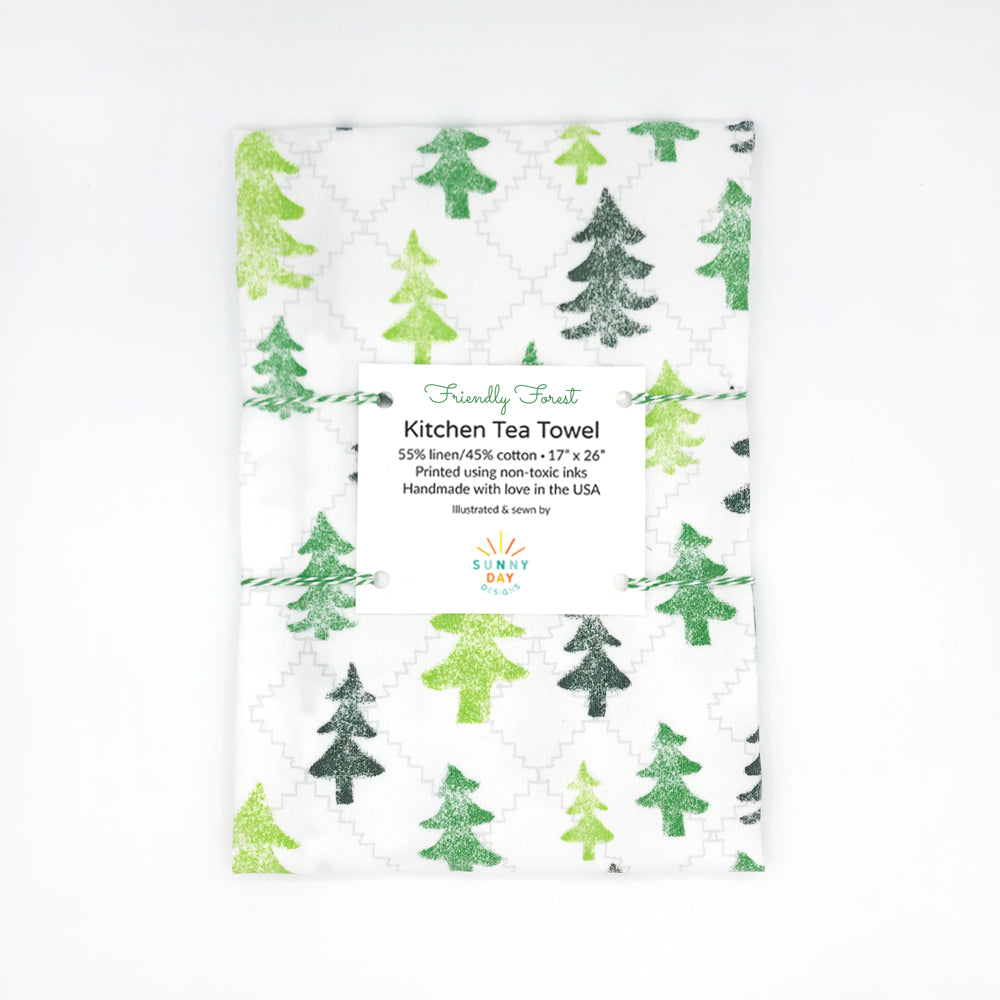 This green Friendly Forest woodland Christmas tree tea towel is handmade from linen/cotton fabric. This green and white Christmas tea towel is folded and packaged on a white background. Made in the USA by Sunny Day Designs