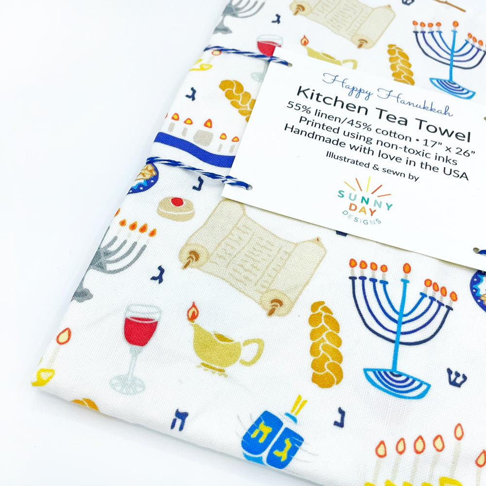 A close up view of the corner of the Happy Hanukkah printed tea towel by Sunny Day Designs. This fun tea towel is a great Jewish gift for Chanukah since it is made in the USA and features illustrated designs of all your Hanukkah favorites: hanukkiah menorahs, challah brad, dreidels, sufganiyot, potato latkes, the torah, and more!