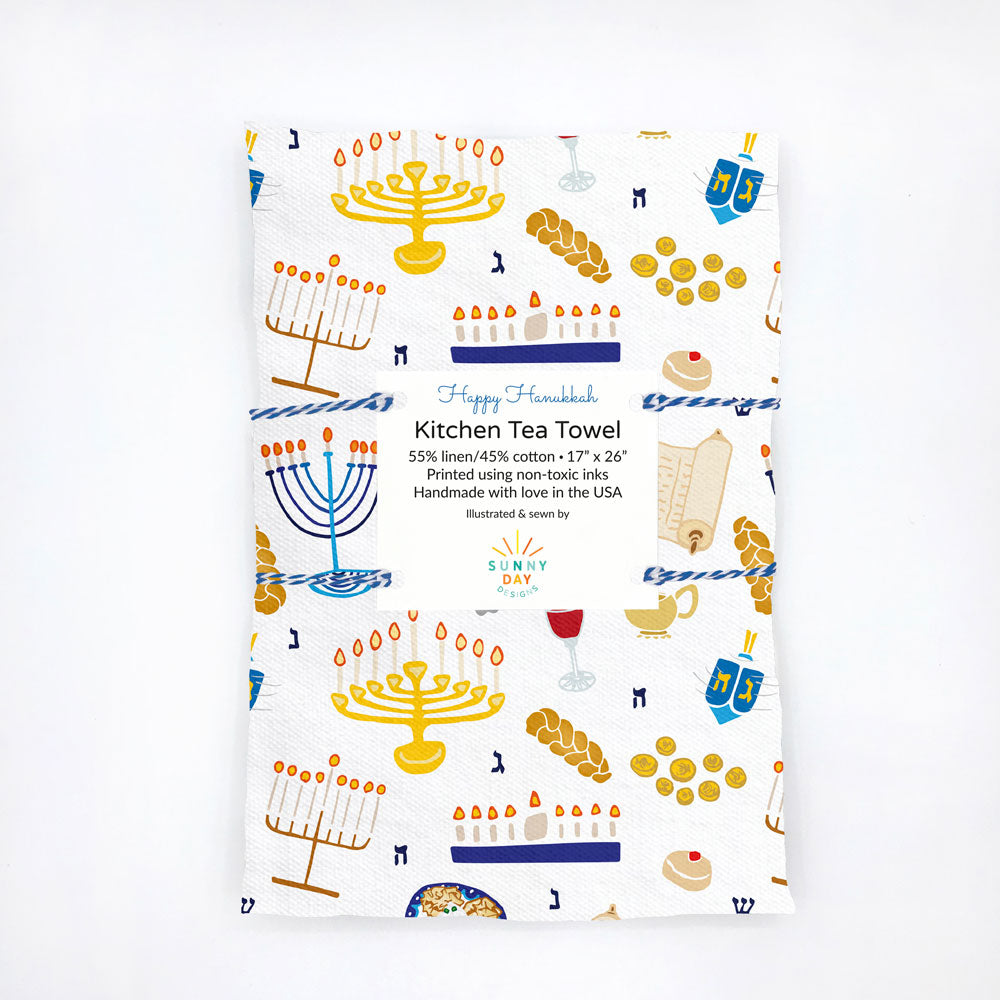 Happy Hanukkah printed kitchen tea towel by Sunny Day Designs is packaged using blue baker's twine and folded on a white background. Each fun dish towel is made in the USA & features illustrated menorahs, dreidels, latkes, challah, and more Chanukah favorites!