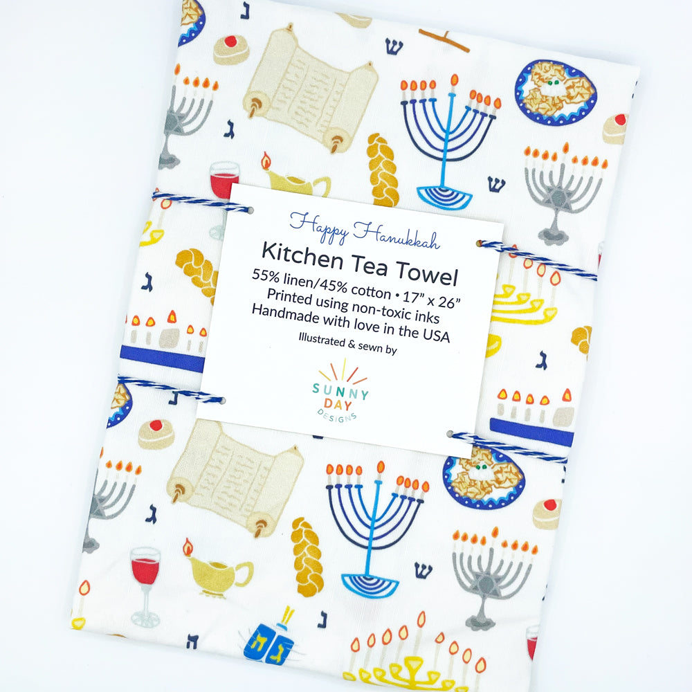 Happy Hanukkah printed tea towel design by Sunny Day Designs. Folded and packaged on a white background. Featuring challah, menorahs, latkes, torah, dreidel, and more Jewish Chanukah favorites! Handmade in the USA.