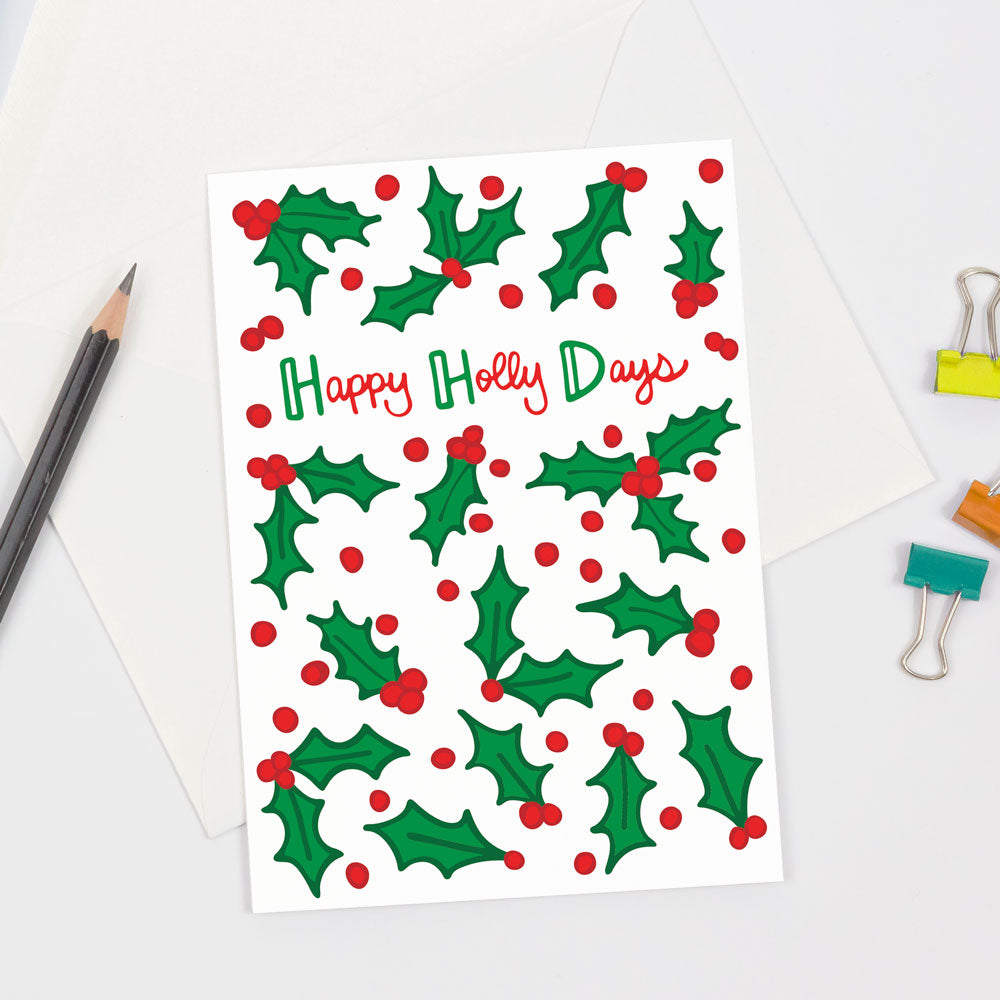 Happy Holly Days Christmas Greeting Card by Sunny Day Designs. Holiday Card with red and green holly berries and leaves on White background. Hand lettered "Happy Holly Days" text on front of card. Sustainably sourced card stock. Made in the USA.
