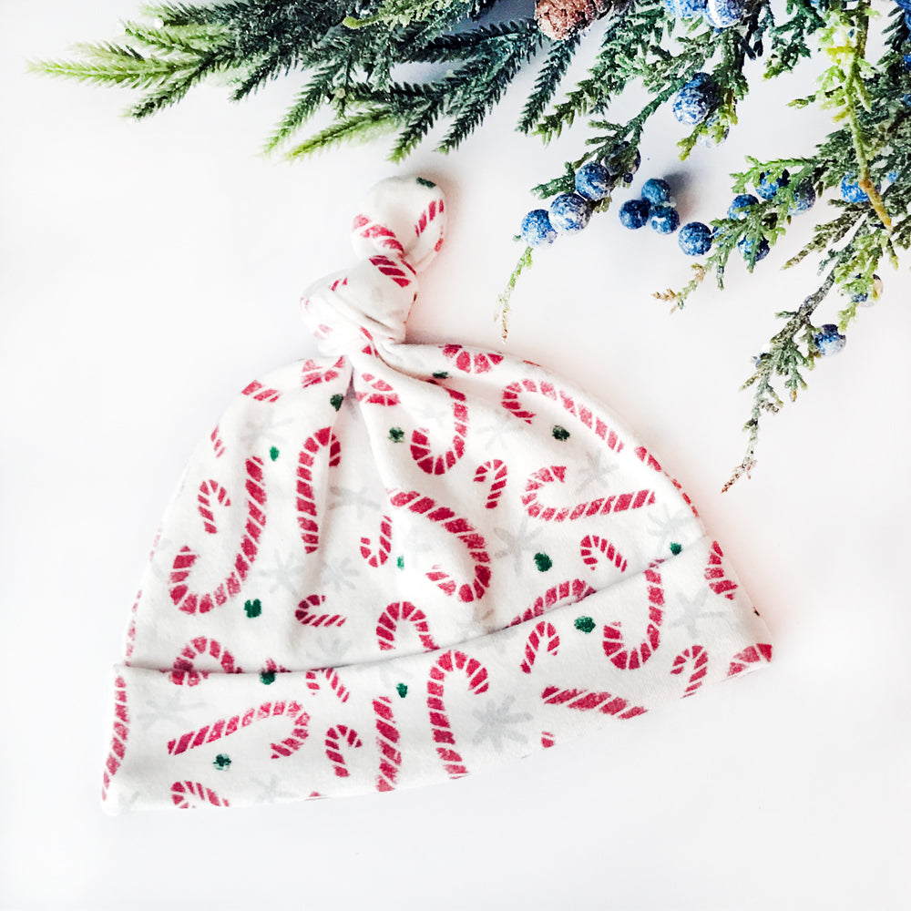 Candy Cane printed organic cotton baby hat by Sunny Day Designs featuring a red candy cane printed newborn hat with a tied top style on a white background with holiday branches and blue juniper berries at the top.
