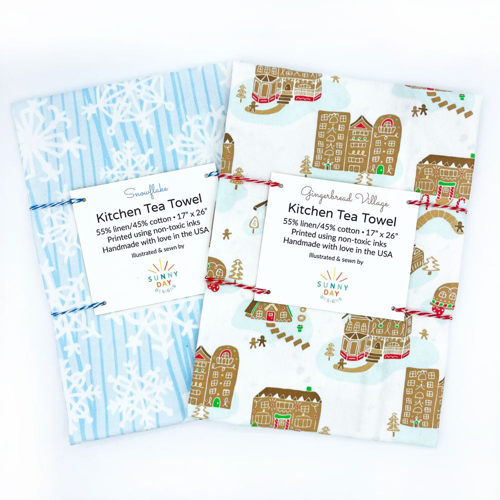 This fun set of 2 handmade holiday tea towels by Sunny Day Designs features 1 blue/white Snowflake winter printed dish towel & 1 whimsical Gingerbread Village printed kitchen towel. Made in the USA & shown folded & packaged on a white background.