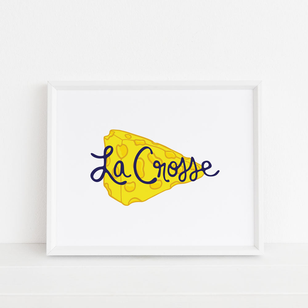 This fun yellow and navy blue La Crosse Wisconsin art print features a yellow wedge of cheese with hand lettered navy blue "La Crosse" text on a white background. Shown framed in a white frame. Printed on watercolor paper in the USA and designed by Sunny Day Designs.