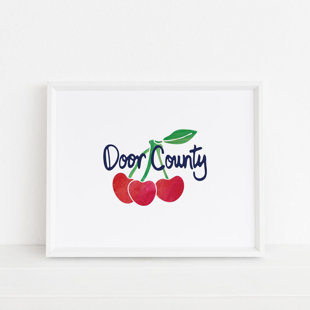 Door County Cherry wall art by Sunny Day Designs. Red and green watercolor cherries on a white background with navy blue handlettered "Door County" text is digitally printed onto white watercolor paper. 8x10 Art Print is shown inside a white frame on a white shelf. This Wisconsin themed wall art is made in the USA.