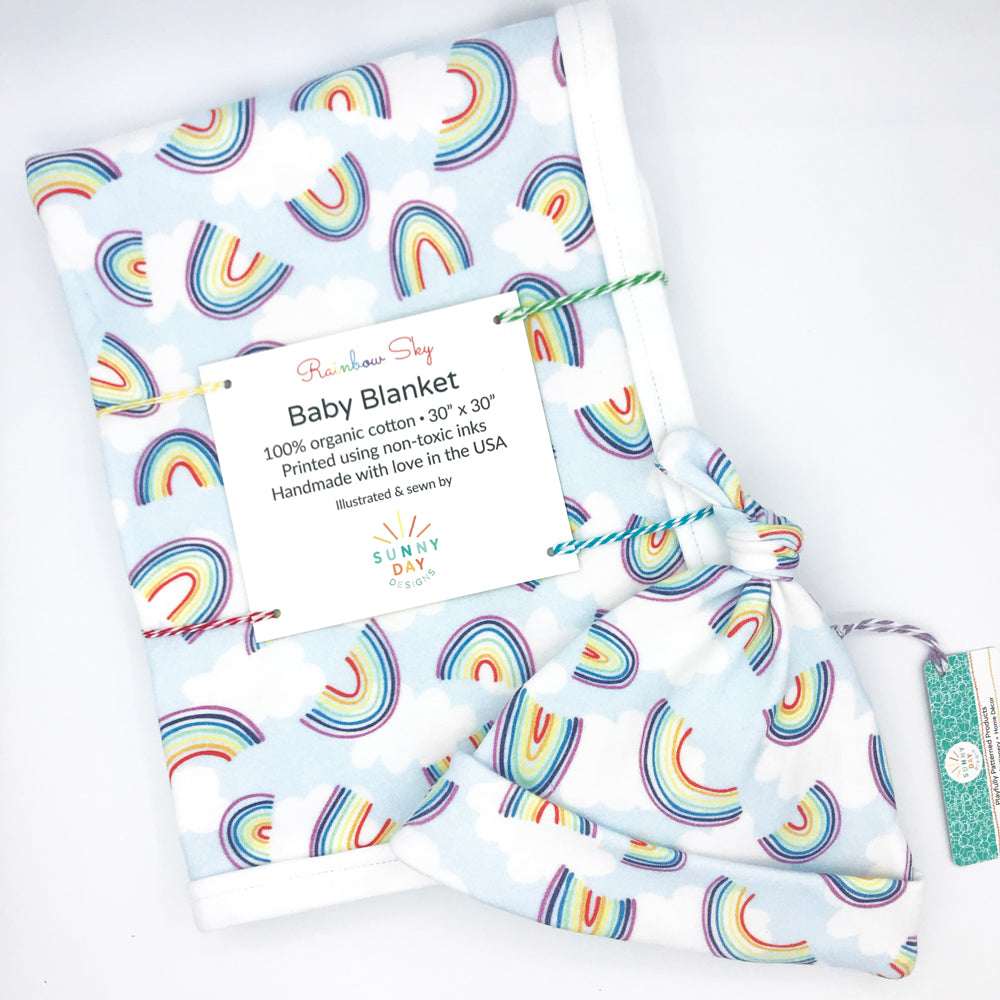 rainbow sky organic cotton baby gift set made in the USA by Sunny Day Designs with newborn hat and baby blanket in multi-color rainbow print design with white clouds on light blue background 