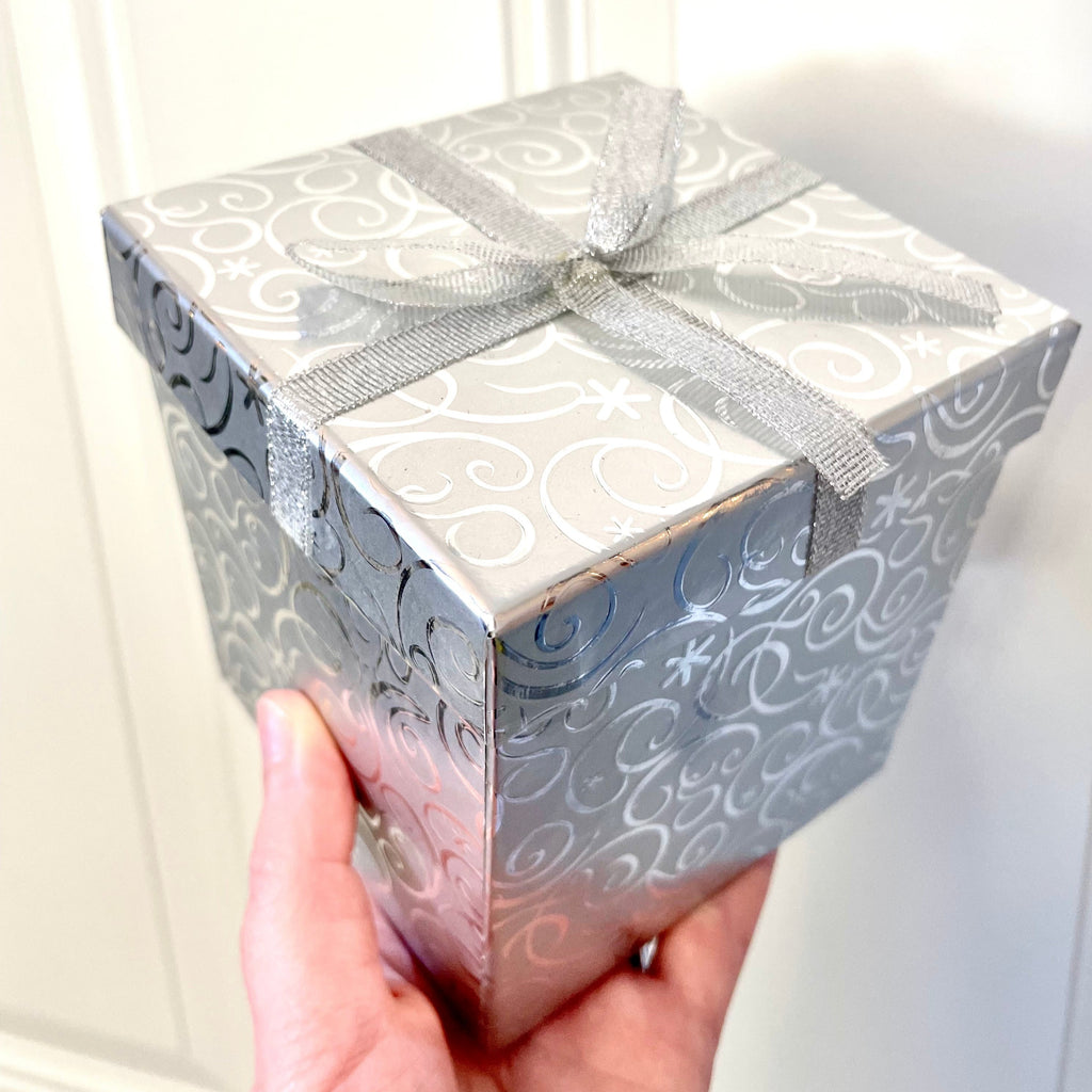 A hand holding a decorative silver gift box in front of a white background, complimentary gift box with each holiday ornament purchase from Sunny Day Designs