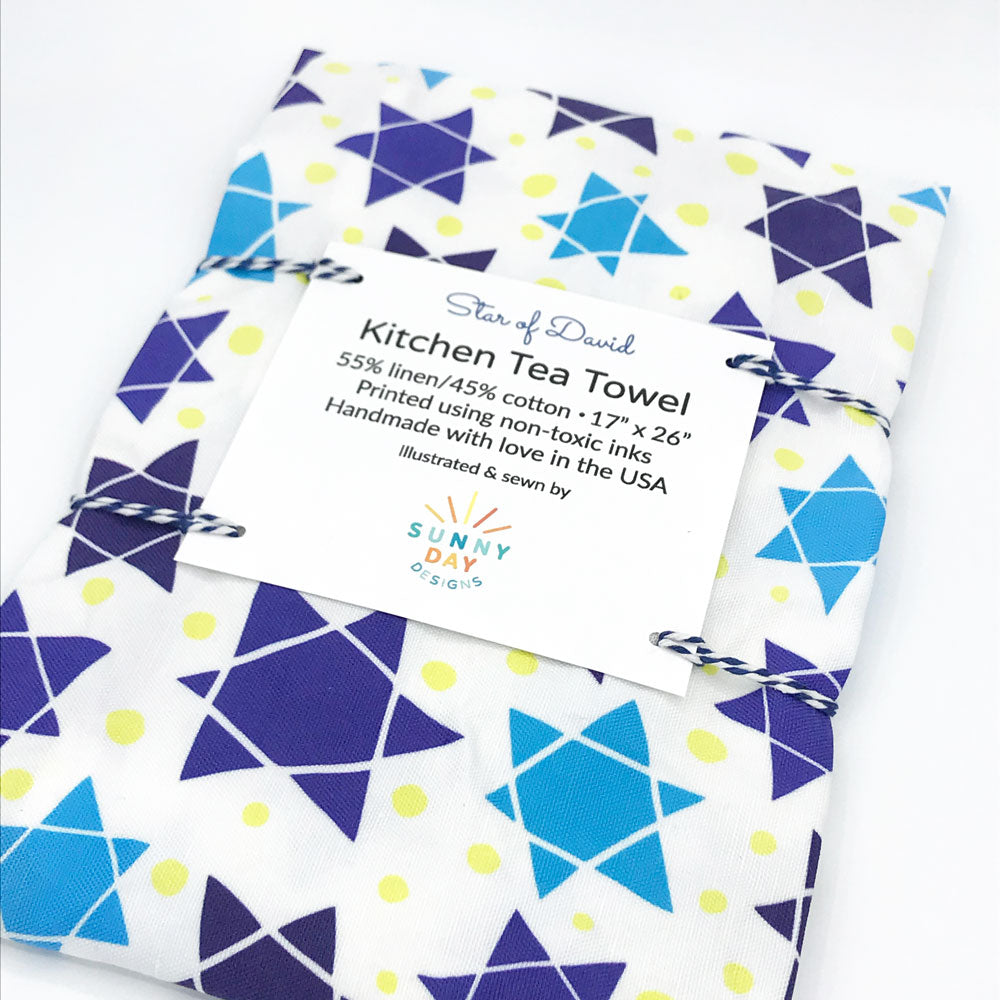 Star of David printed fun tea towel by Sunny Day Designs featuring abstract star of david printed symbols in various shades of blue on a white background with yellow dots. This handmade tea towel is made in the USA and folded/packaged with blue striped baker's twine.