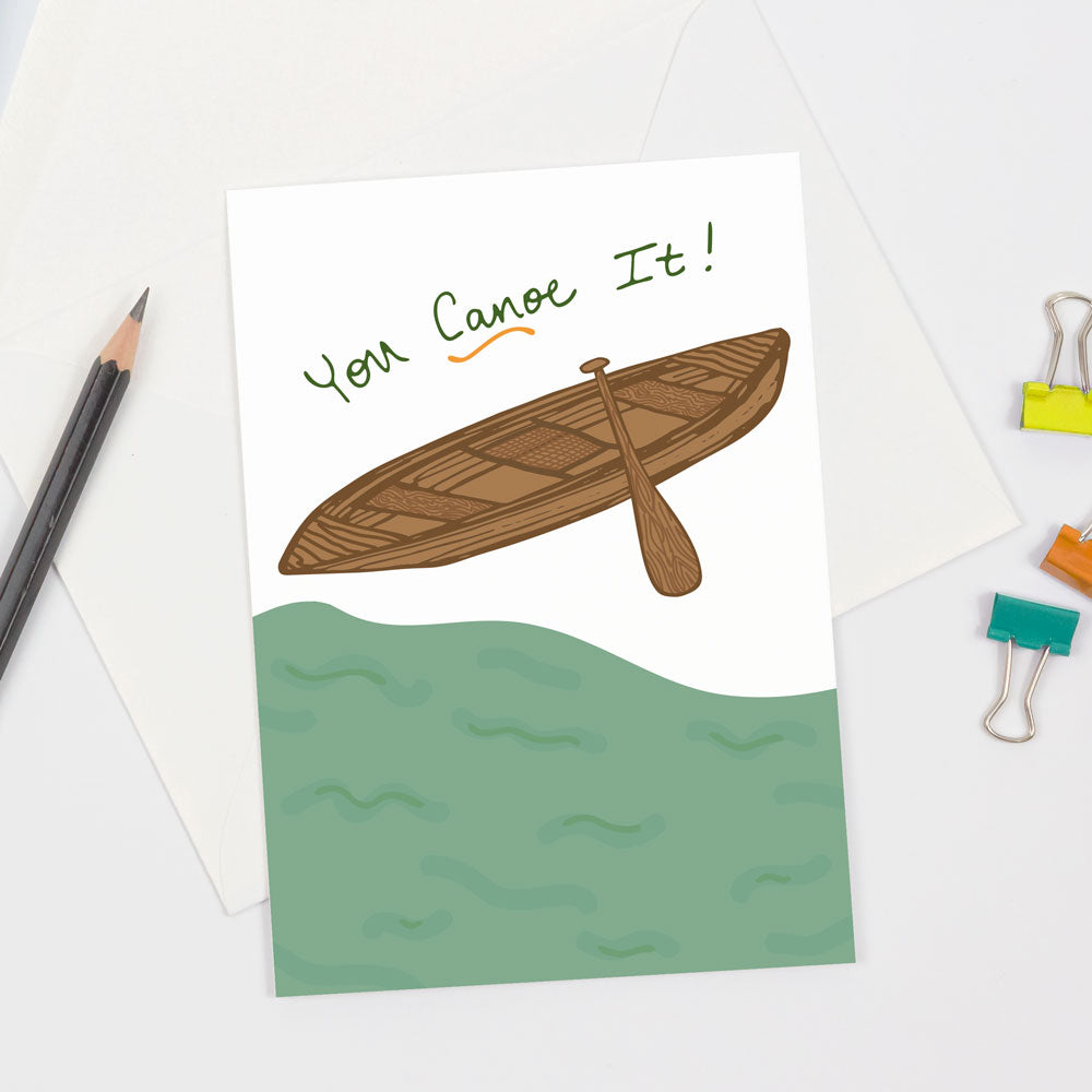 An encouragement greeting card with humorous text that says "You Canoe It!" and an illustrated brown wooden canoe and 1 wooden paddle resting on the bank of a green lake. This card is designed by Sunny Day Designs and is digitally printed and made in the USA. Shown on a white table alongside a sharpened pencil and 3 colorful binder clips.